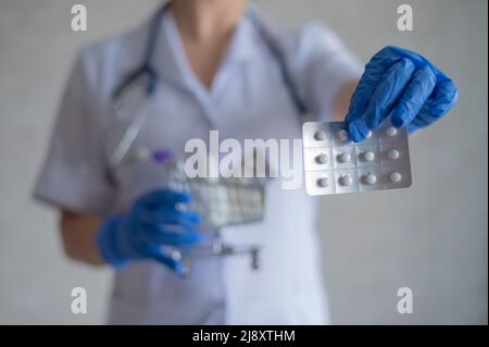 Un medico senza volto in guanti di gomma mostra un blister con pillole e contiene un mini carrello con una confezione completa di diversi farmaci Foto Stock