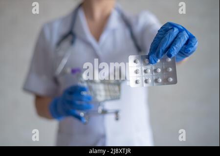 Un medico senza volto in guanti di gomma mostra un blister con pillole e contiene un mini carrello con una confezione completa di diversi farmaci Foto Stock