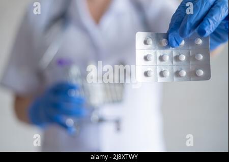 Un medico senza volto in guanti di gomma mostra un blister con pillole e contiene un mini carrello con una confezione completa di diversi farmaci Foto Stock