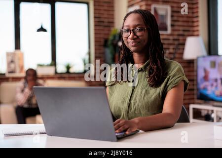 Ritratto di imprenditore afroamericano che lavora lontano da casa sorridendo a macchina fotografica mentre il ragazzo si sta rilassando sul divano. Donna casual con occhiali in posa davanti al computer portatile a casa. Foto Stock