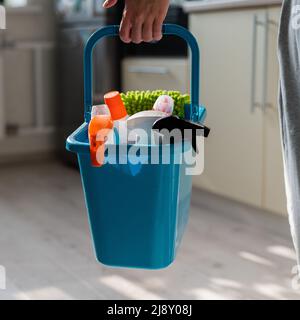 Concetto di pulizia della casa. La donna senza volto contiene i prodotti per la pulizia in un cestino blu. Primo piano delle mani di una casa operaia femminile. Foto Stock