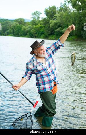 Pescatore che pesca su un rever. Un vecchio pescatore felice ha preso un pesce di trota. Ritratto di allegro sorridente uomo anziano pesca. Nonno con pesce di cattura. Foto Stock