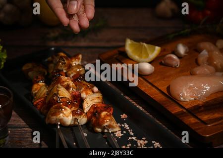 Uomo cospargere a mano sale e condimento su spiedini di pollo e verdure alla griglia Foto Stock