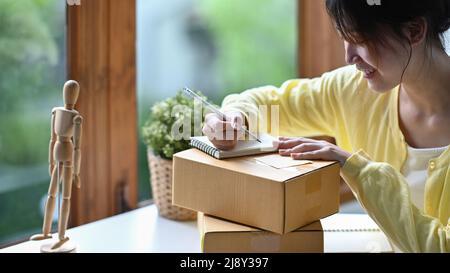 Indirizzo di scrittura del venditore in linea della giovane donna messo a fuoco sulla scatola di cartone mentre preparando le scatole del pacco del prodotto per la consegna Foto Stock