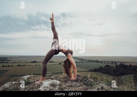 Giovane donna che fa esercizi ginnici sulla cima di una collina all'aperto, bellissimo paesaggio sullo sfondo Foto Stock