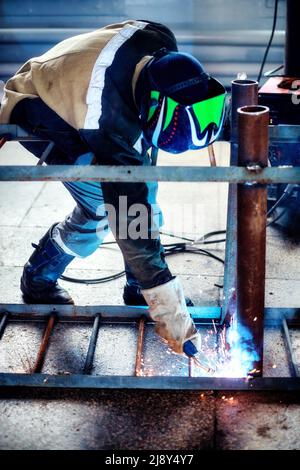 Saldatrice professionale in uniforme protettiva e maschere in sala di produzione. Flusso di lavoro autentico. La saldatrice lavora con metallo e scintille volare. Foto Stock