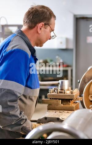 Un anziano tornitore di 50-55 anni in tute lavora al tornio in produzione. Lavorazione di prodotti metallici. Ritratto reale di lavoratore caucasico. Background di produzione. Foto Stock