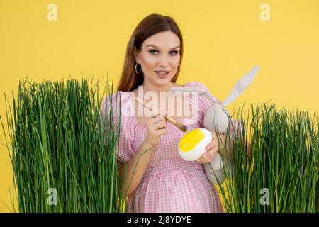 Donna in tarda gravidanza su decorazione erba dipingere uovo con spazzola e tenere giocattolo morbido coniglio, sfondo giallo. Ritratto di giovane donna incinta Foto Stock