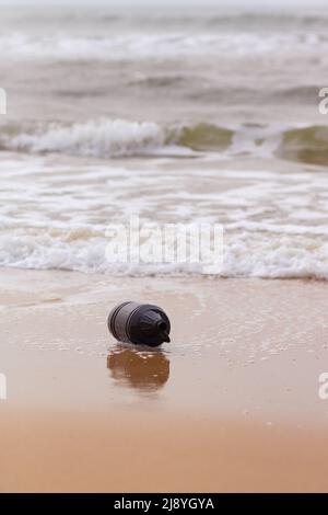 Una bottiglia vuota di plastica nera sulla spiaggia che viene trascinata dalle onde dell'oceano sulla sabbia. Concetto di inquinamento dell'acqua e del mare. Composizione verticale Foto Stock