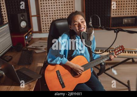 Il giovane musicista africano canta nel microfono mentre suona la nuova canzone e suona la chitarra nello studio di registrazione Foto Stock