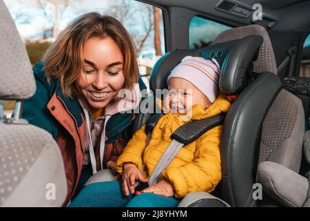 Una mamma caucasica sorridente sta preparando il suo piccolo bambino grazioso per un viaggio in auto. Una donna allaccia le cinture di sicurezza in un seggiolino per bambini nello schienale. Visualizza f Foto Stock