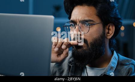 Messo a fuoco indiano barbuto uomo freelancer in occhiali che lavorano sul computer a casa tarda notte guardando la decisione di ricerca schermo si sente dubbio cr Foto Stock