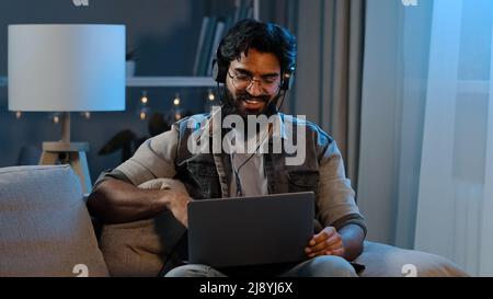 L'uomo bearded divertente indiano arabo indossa cuffie senza fili che hanno divertimento ascoltare la musica con il laptop a casa seduta divano sera tardi Darkne tempo Foto Stock