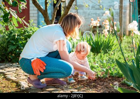 La giovane madre insegna alla figlia del bambino a curare i letti di erbe con una pala giocattolo. Vista laterale. Famiglia estate giardinaggio concetto. Foto Stock