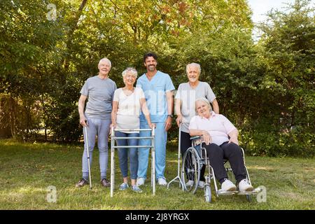 Gruppo di anziani con camminatori e sedie a rotelle nel giardino della clinica di riabilitazione insieme ad un'infermiera Foto Stock