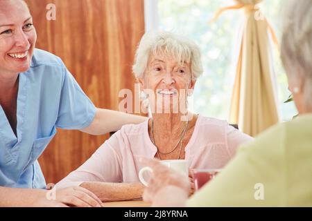 Uomo anziano e caregiver sorridente che parlano mentre si prende il caffè in casa di cura Foto Stock