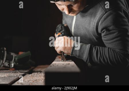 Un uomo lavora con un trapano nella sua officina. Trapani da carpentiere con utensile elettrico a mano in una stanza buia con luce direzionale Foto Stock