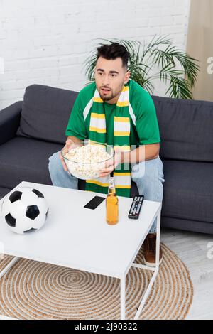 sorprende il tifoso dello sport con una ciotola di popcorn che guarda la partita in tv vicino alla birra e alla palla di calcio Foto Stock