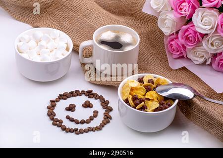 Buona colazione. Cereali con latte, caffè con marshmallows, un bouquet di rose e un sorriso sorridente fatto di chicchi di caffè Foto Stock