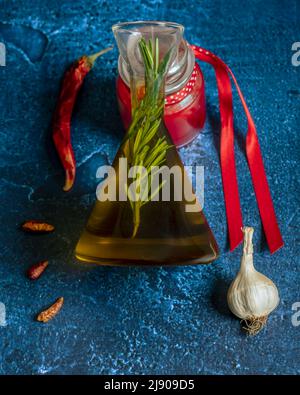 Un crudo di olio extravergine di oliva con un rametto di rosmarino all'interno e aglio e peperoncino su sfondo blu Foto Stock