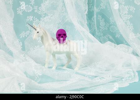 un unicorno bianco lucido che porta un cranio di plastica sul dorso. Fotografia a colori a vita fissa minimale e creativa Foto Stock
