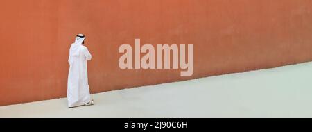 Banner orizzontale con uomo arabo con abiti tradizionali emirates che camminano all'aperto in strada contro un muro di sfondo arancione - etnia araba a Foto Stock