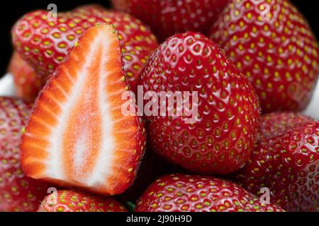 Primo piano di fragole rosse fresche mature senza code verdi su sfondo nero. Una fragola tagliata a metà tra fragole intere Foto Stock