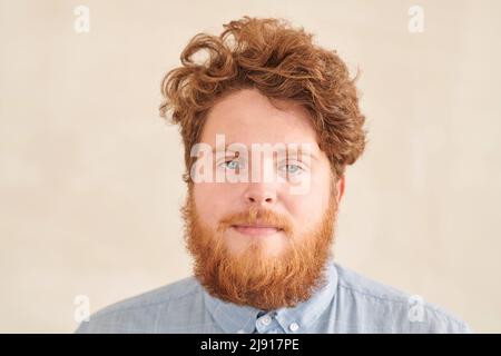 Ritratto di giovane studente rosso serio con barba e baffi con capelli ricci in piedi su sfondo beige Foto Stock