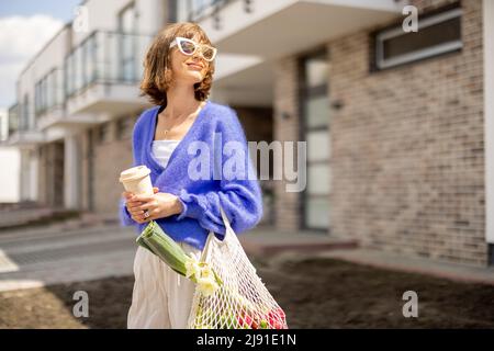 Donna cammina con sacco a rete pieno di verdure e tazza di caffè riutilizzabile vicino a casa all'aperto Foto Stock