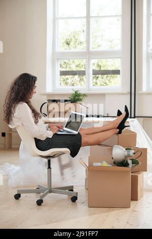 Donna d'affari giovane e positiva in gonna seduta con piedi su una pila di scatole mobili e lavorare con il laptop in un nuovo ufficio Foto Stock