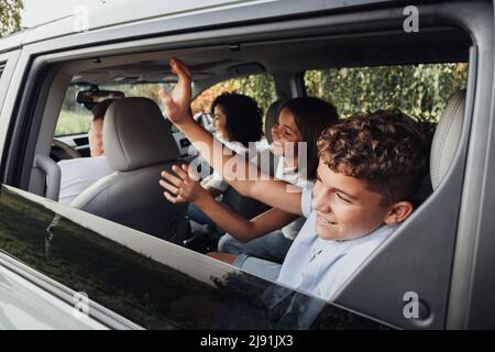 Adolescente ragazzo e ragazza saluto a qualcuno attraverso la finestra con le mani ondeggianti mentre si siede all'interno di minivan auto, felice quattro membri Famiglia in un fine settimana Foto Stock