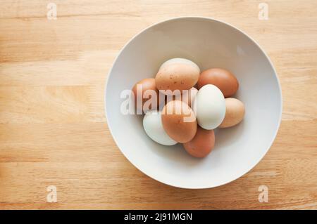 Uova fresche in tonalità di marrone e bianco in una ciotola bianca su tavola di legno, immagine presa dall'alto, con spazio copia. Foto Stock