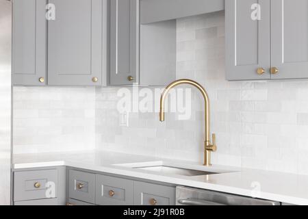 Un dettaglio lavandino della cucina girato con un rubinetto d'oro, backsplash in marmo, armadi grigi, e metallo dorato. Foto Stock