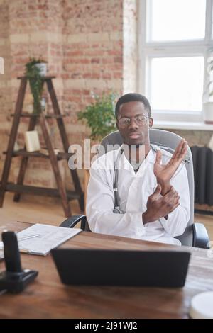 Ritratto del medico africano americano moderno seduto in camera d'ufficio loft controllo della sua frequenza cardiaca su macchina fotografica, concetto di telemedicina Foto Stock