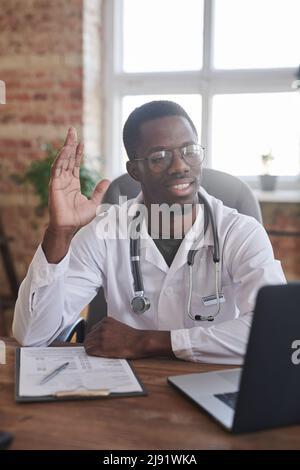 Bel medico afroamericano che indossa cappotto bianco e occhiali seduti alla scrivania di fronte al computer portatile in camera d'ufficio loft saluto il suo paziente online Foto Stock