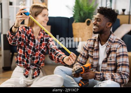 Giovane donna con metro a nastro che guarda l'uomo sorridente con trapano elettrico mentre si siede insieme sul pavimento. Coppia multirazziale che misura lo spazio libero per nuovi mobili in appartamento moderno. Foto Stock