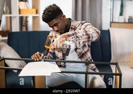 Ritratto di felice uomo afroamericano in abbigliamento casual con avvitatore a batteria in mano che monta armadio moderno in soggiorno. Un nuovo concetto di casa. Foto Stock