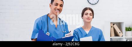 Allegri veterinari interrazziali con appunti e tablet digitale guardando la fotocamera in clinica, banner Foto Stock