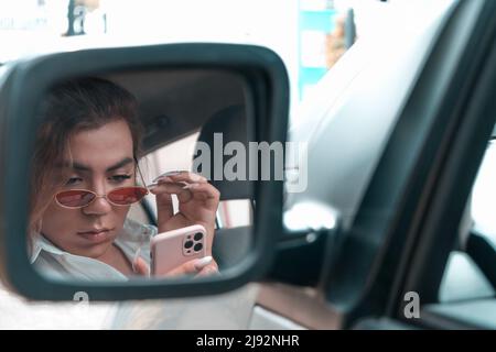 giovane uomo androgino che guarda sorpreso e che legge il suo telefono in auto nello specchio retrovisore. Foto di alta qualità Foto Stock