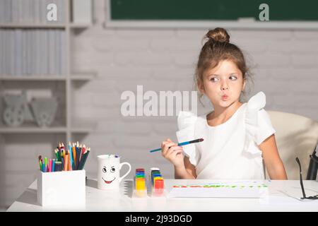 Bambina attira in aula seduto a un tavolo, divertendosi sulla lavagna di fondo della scuola. Apprendimento infantile, abilità artistiche dei bambini. Foto Stock