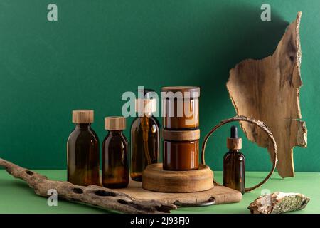 Contenitori in vetro marrone per cosmetici con coperchi in legno. Vasetto crema, flacone contagocce e lozione. Sfondo verde. Cura e bellezza. Bottiglie. Concetto ecologico. Casella Set cosmetico. Pipetta e dispenser. Foto Stock