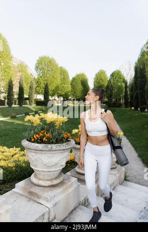 donna in abbigliamento sportivo bianco che cammina con tappetino per il fitness sulle scale del parco Foto Stock