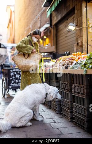Donna in maschera medica acquisto frutta e verdura con cane alla bancarella del mercato sulla famosa strada gastronomica di Bologna. Concetto di acquisto locale pro Foto Stock