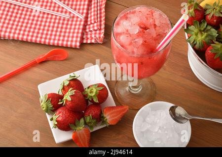 Granita di fragole con frutta matura intorno su tavola di legno con ciotola con ghiaccio e cestino pieno di fragole. Vista dall'alto. Composizione orizzontale Foto Stock