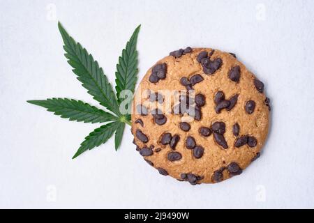 Biscotti al cioccolato con estratto di cannabis in primo piano. Dolci con olio CBD. Foglia di marijuana su sfondo bianco. Vista dall'alto, piatto. Foto Stock