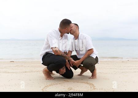 Coppia gay baciare accanto ad un cuore disegnato nella sabbia su una spiaggia tropicale Foto Stock