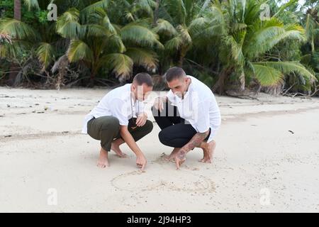 Coppia omosessuale maschile che attingendo un cuore nella sabbia su una spiaggia tropicale Foto Stock