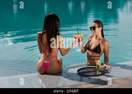 Due bellissime felicemente donne in bikini che tengono bicchieri di cocktail fresco da sorseggiare presso il pool resort. Due eleganti amici femminili tostano con bevande, mentre riposano in piscina. Concetto di celebrazione. Foto Stock