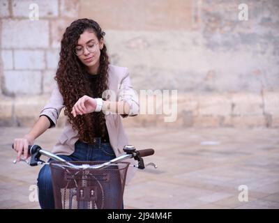 la donna araba che guida un veicolo non inquinante per lavorare in una caffetteria, guarda il tempo sul suo orologio, vista frontale Foto Stock