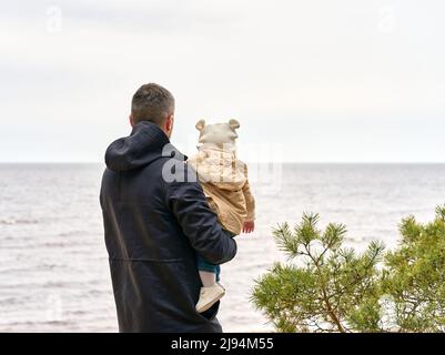 Padre che tiene il bambino sulle sue mani. Guardano all'orizzonte che si erge sulla riva del mare. Pino sul lato destro. Foto Stock